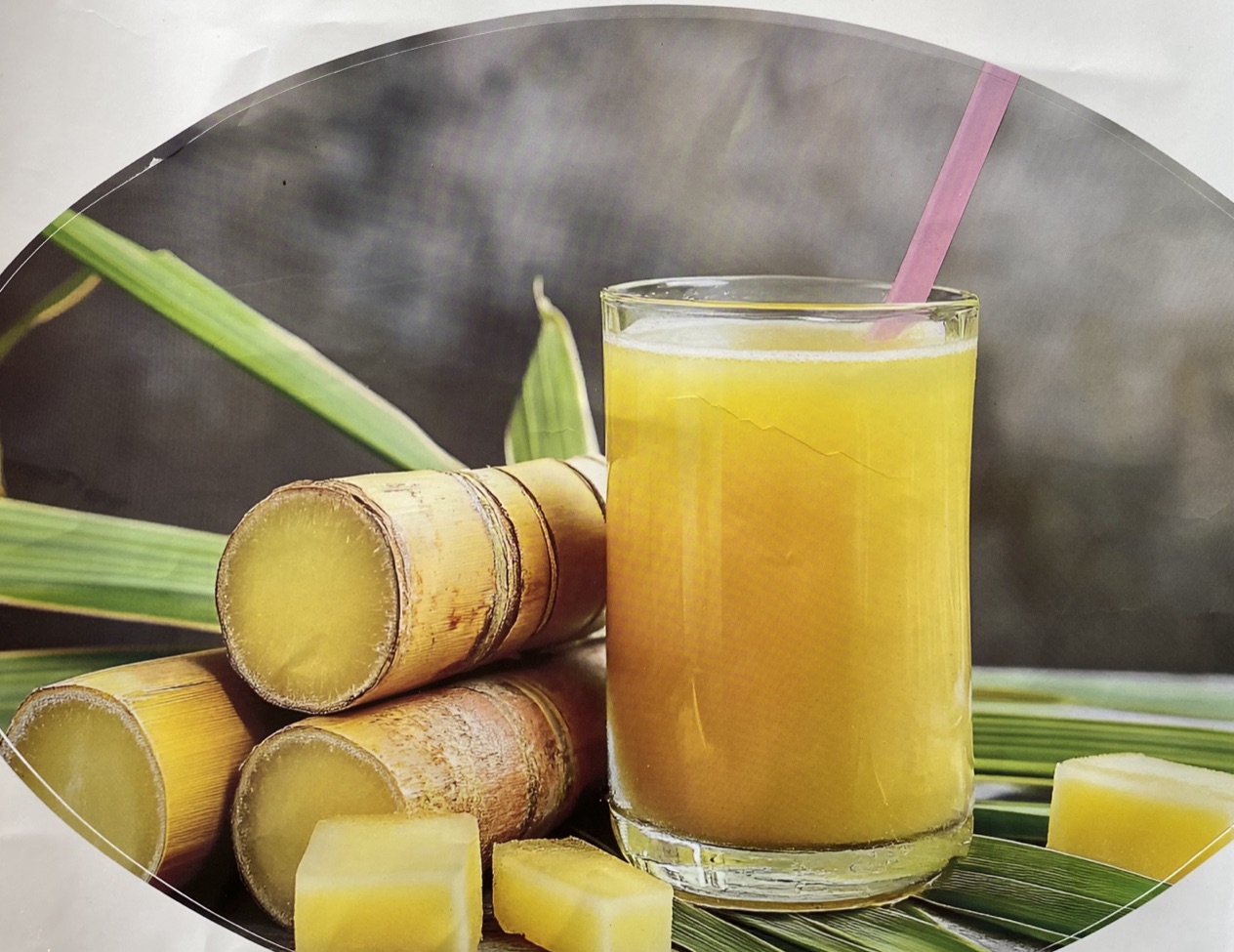 A tall glass of fresh-pressed sugarcane juice (E2 鮮榨蔗汁) with a straw, beside cut sugarcane stalks and cubes